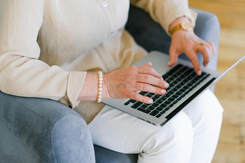 Von oben: Anonyme ältere Frau tippt im Wohnzimmer, während sie in einem Sessel sitzt, Informationen für einen Bericht auf einem Netbook.